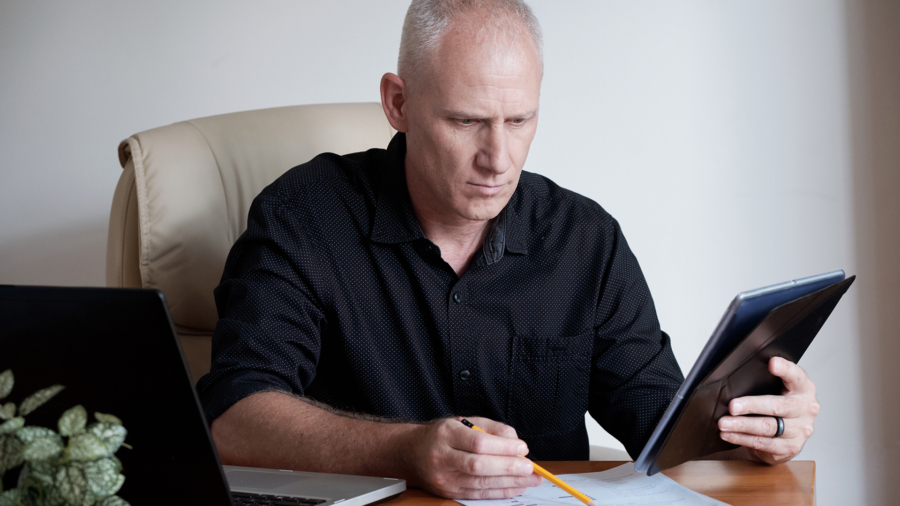 Concentrated mature Caucasian entrepreneur using digital table while sitting at office table and checking data on documents